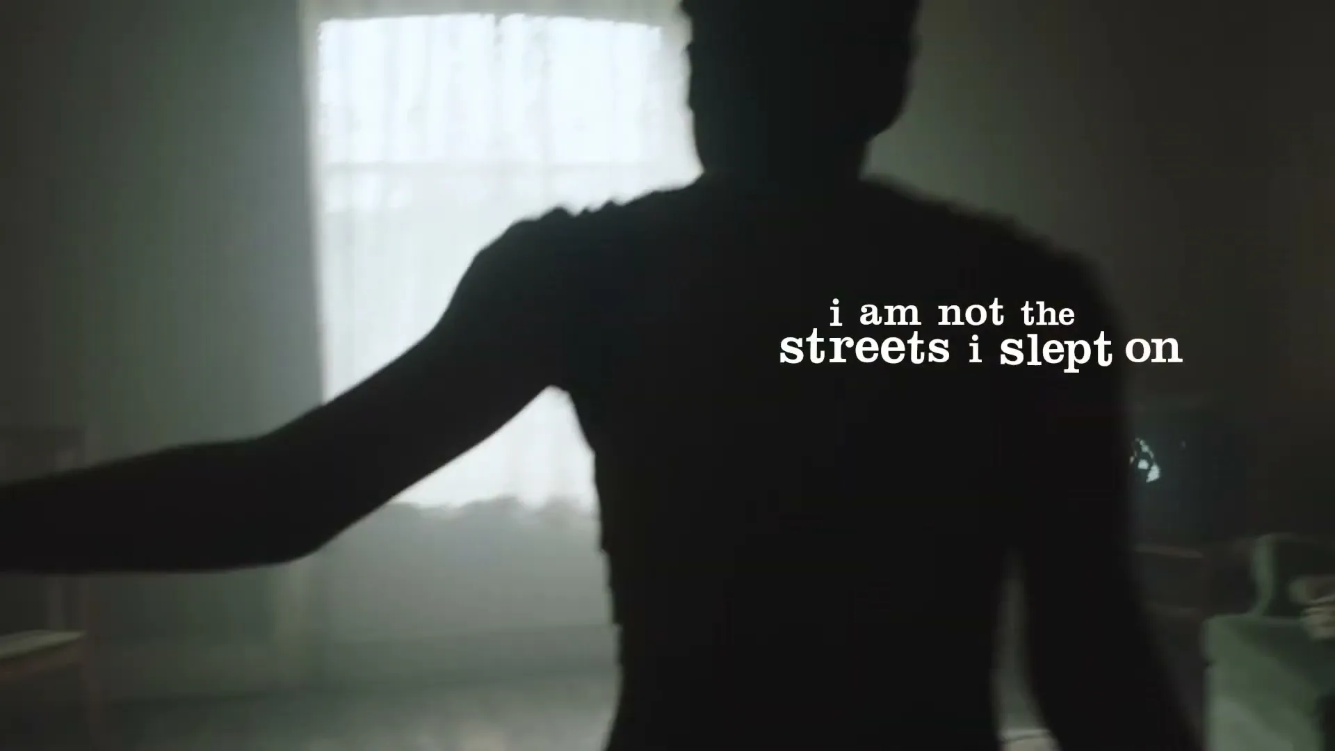 A young person in silhouette dances in a dimly lit room with a window and a television in the background.