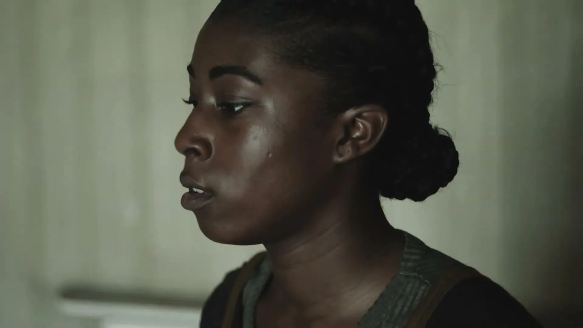 A young Black woman with her hair in a bun looks off-screen, appearing thoughtful or concerned in a dimly lit room.