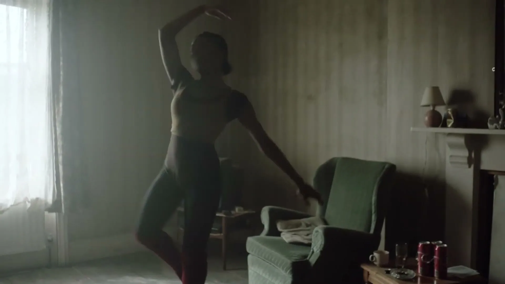 A young Black woman in a leotard practices ballet in a dimly lit, sparsely furnished room with a window and a green armchair.