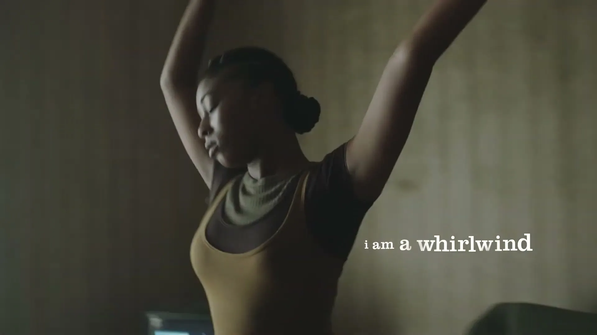 A young Black woman with braided hair, wearing a tank top and leggings, dances in a dimly lit room with a window in the background.
