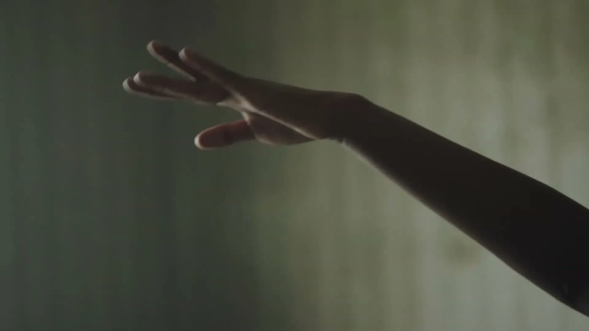 A child's arm and hand, silhouetted against a dimly lit wall, slowly reaches out in a graceful, ballet-like movement.