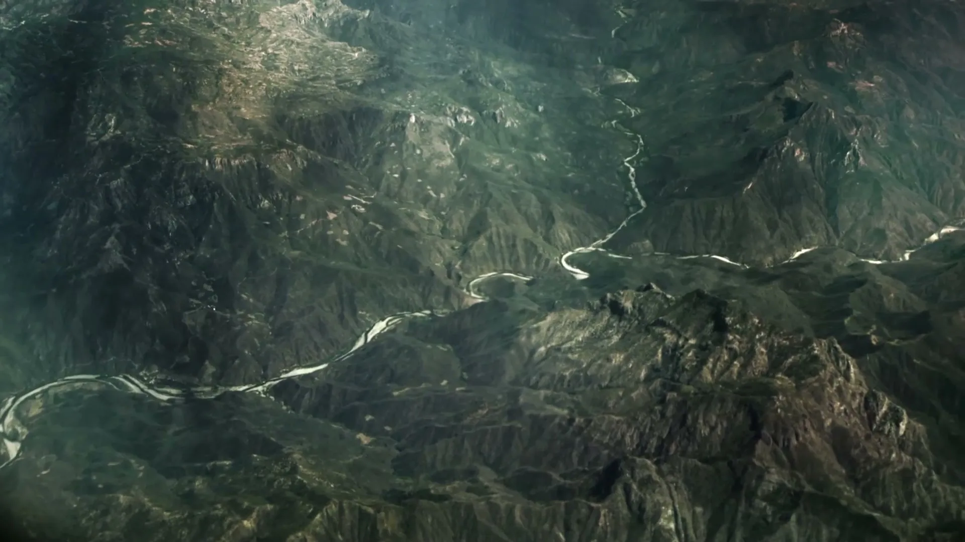 An aerial view shows a winding river cutting through a vast, mountainous landscape under a hazy sky.