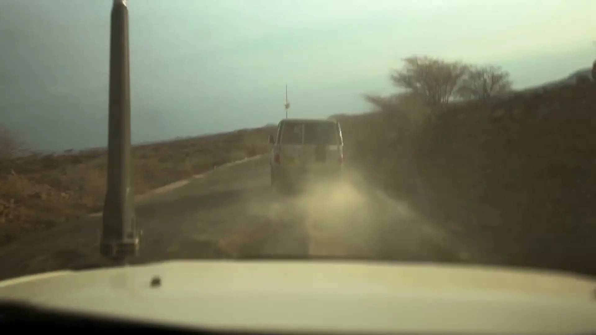 A POV shot from inside a vehicle shows a dusty road with another vehicle driving ahead, kicking up dust in a dry, rural landscape under a hazy sky.