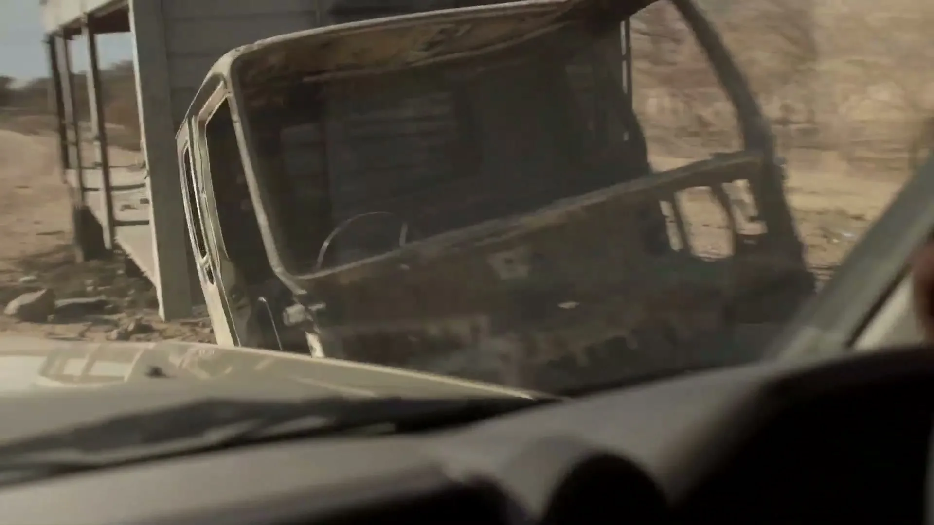 A vehicle drives past a rusted, abandoned truck on a dusty road in a dry, rural landscape.