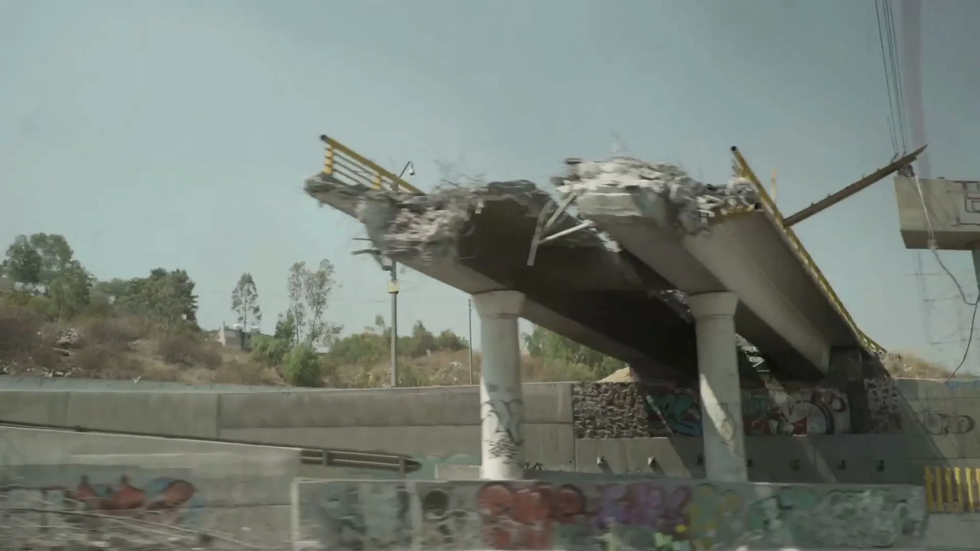 A wide shot of a destroyed concrete overpass with graffiti on its base, under a clear sky, suggesting urban decay or disaster.