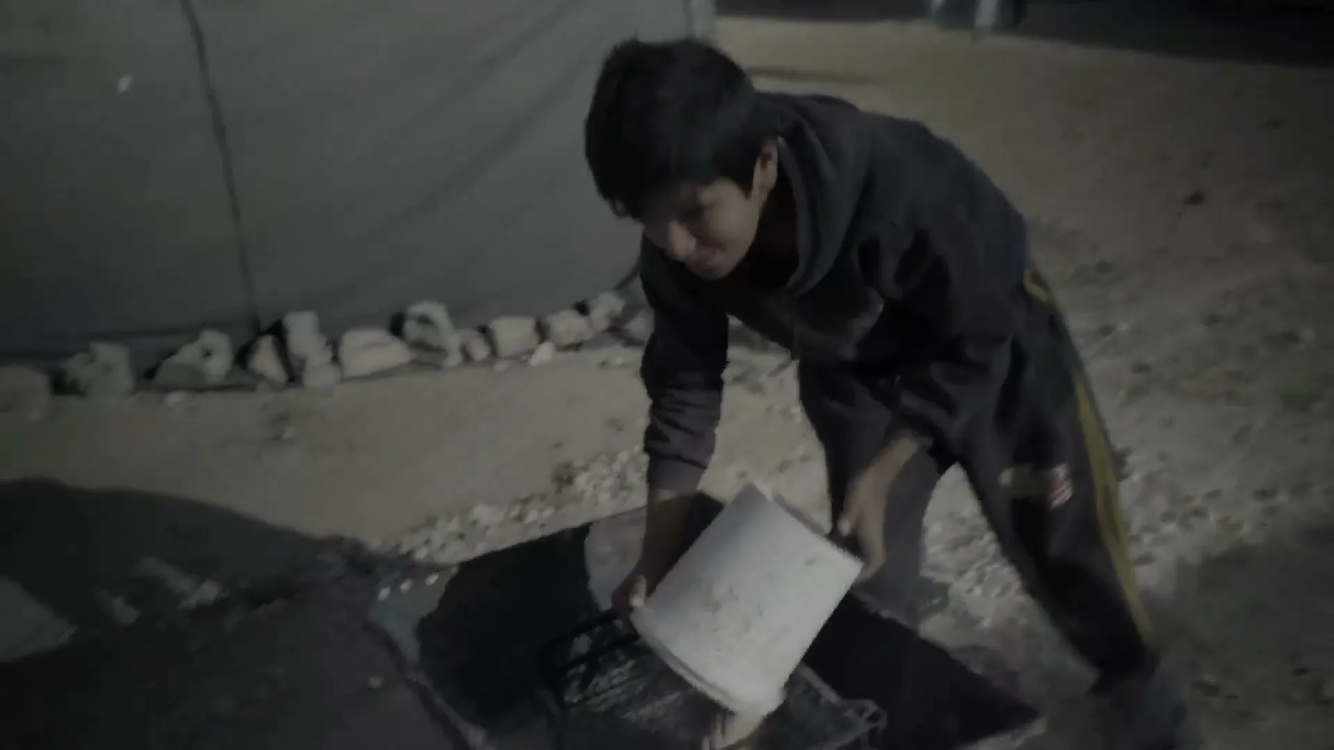 A young boy in a dark hoodie and sweatpants pours water from a white bucket into a metal container on the ground at night in a refugee camp.