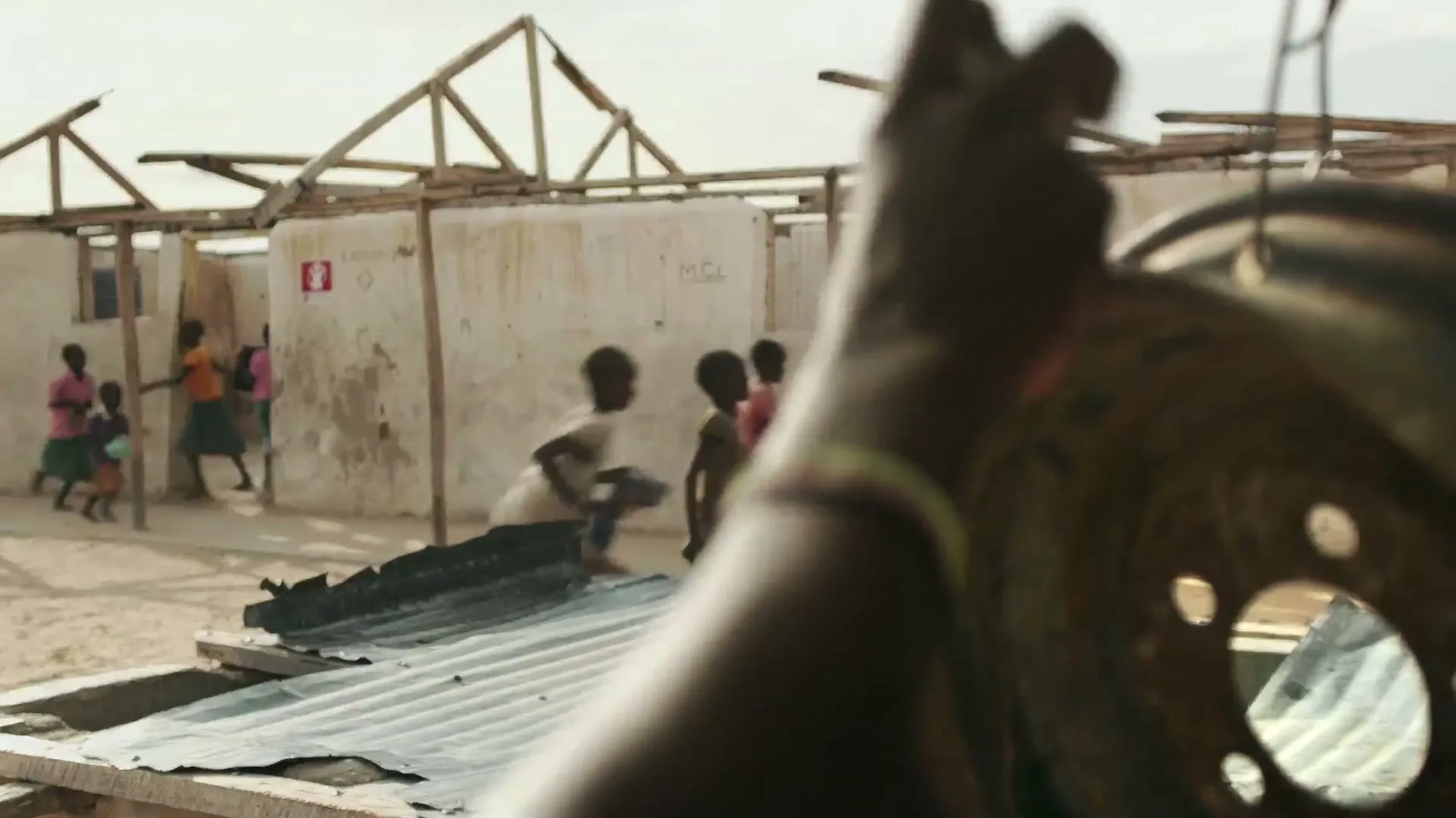 A child's arm with a beaded bracelet holds a metal object in the foreground, while other children play in a dusty, makeshift village in the background.