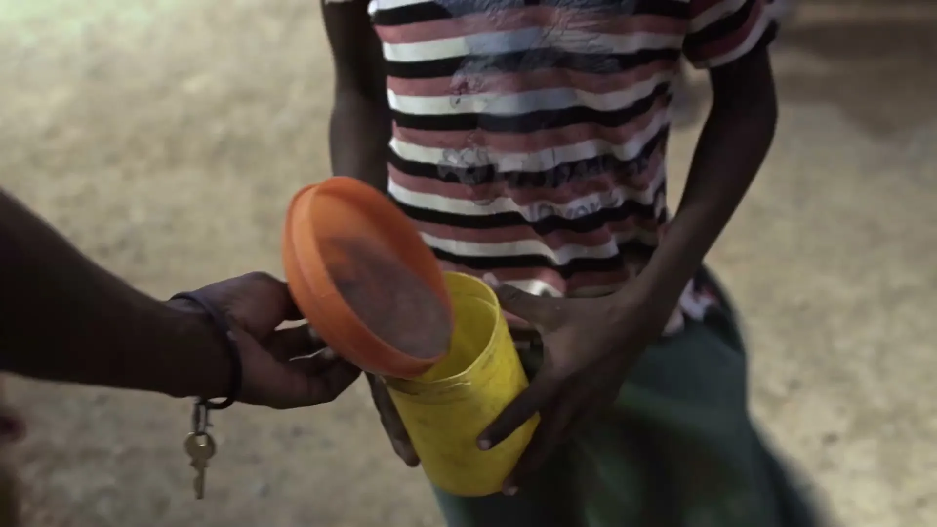 A person pours a small amount of food from an orange lid into a yellow container held by a child in a striped shirt.