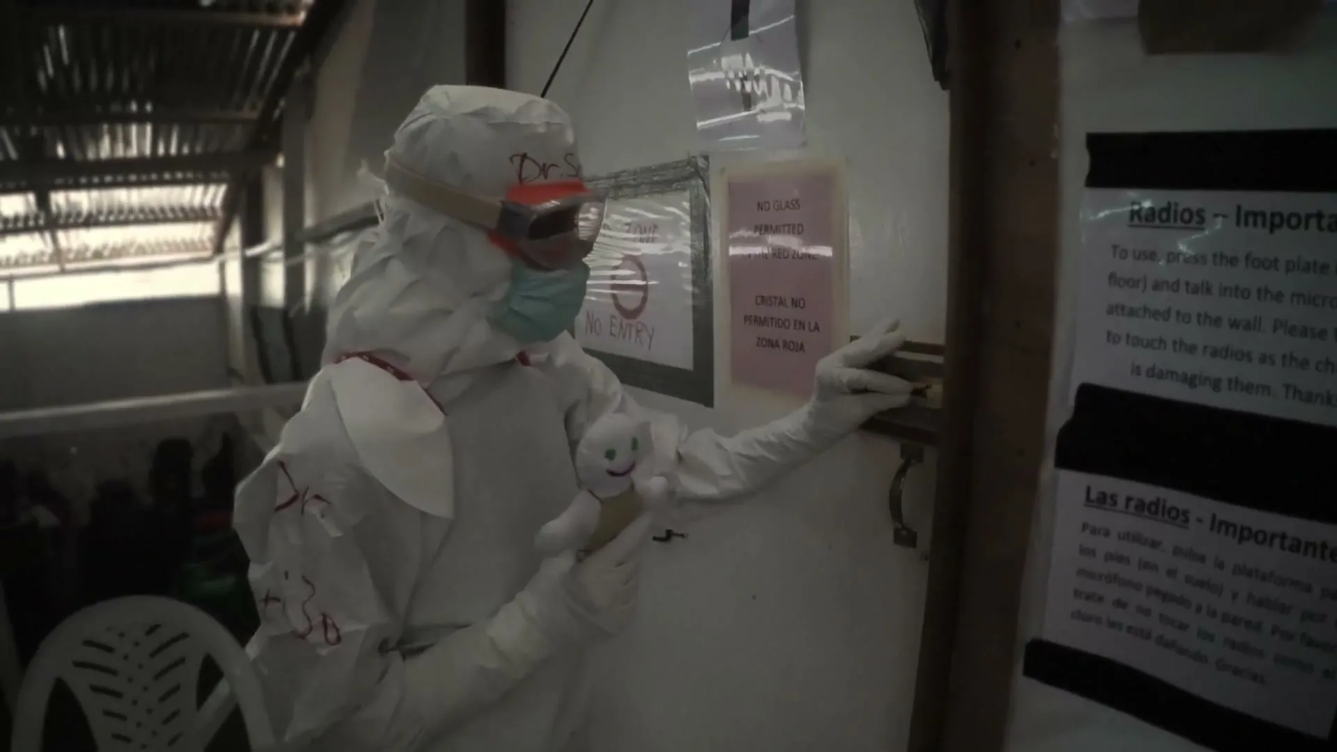A medical worker in a full white hazmat suit, goggles, and a face mask holds a small white doll while standing in a room with signs on the wall.