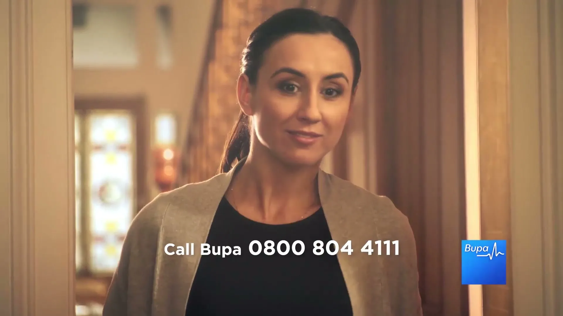 A woman in her 30s, wearing a black top and beige cardigan, smiles gently at the camera in a well-lit home interior.