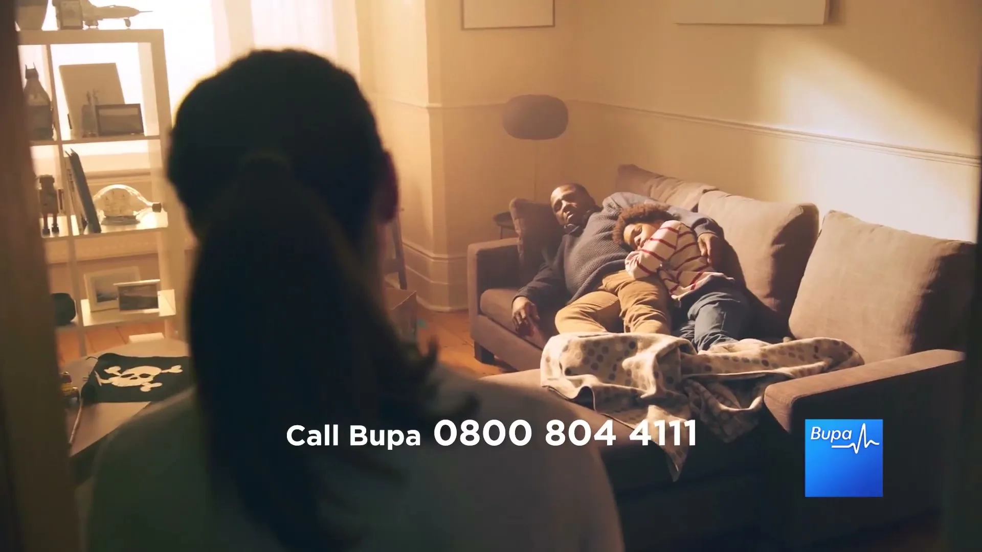 A woman with a ponytail looks into a living room where a man and a child are sleeping on a sofa.