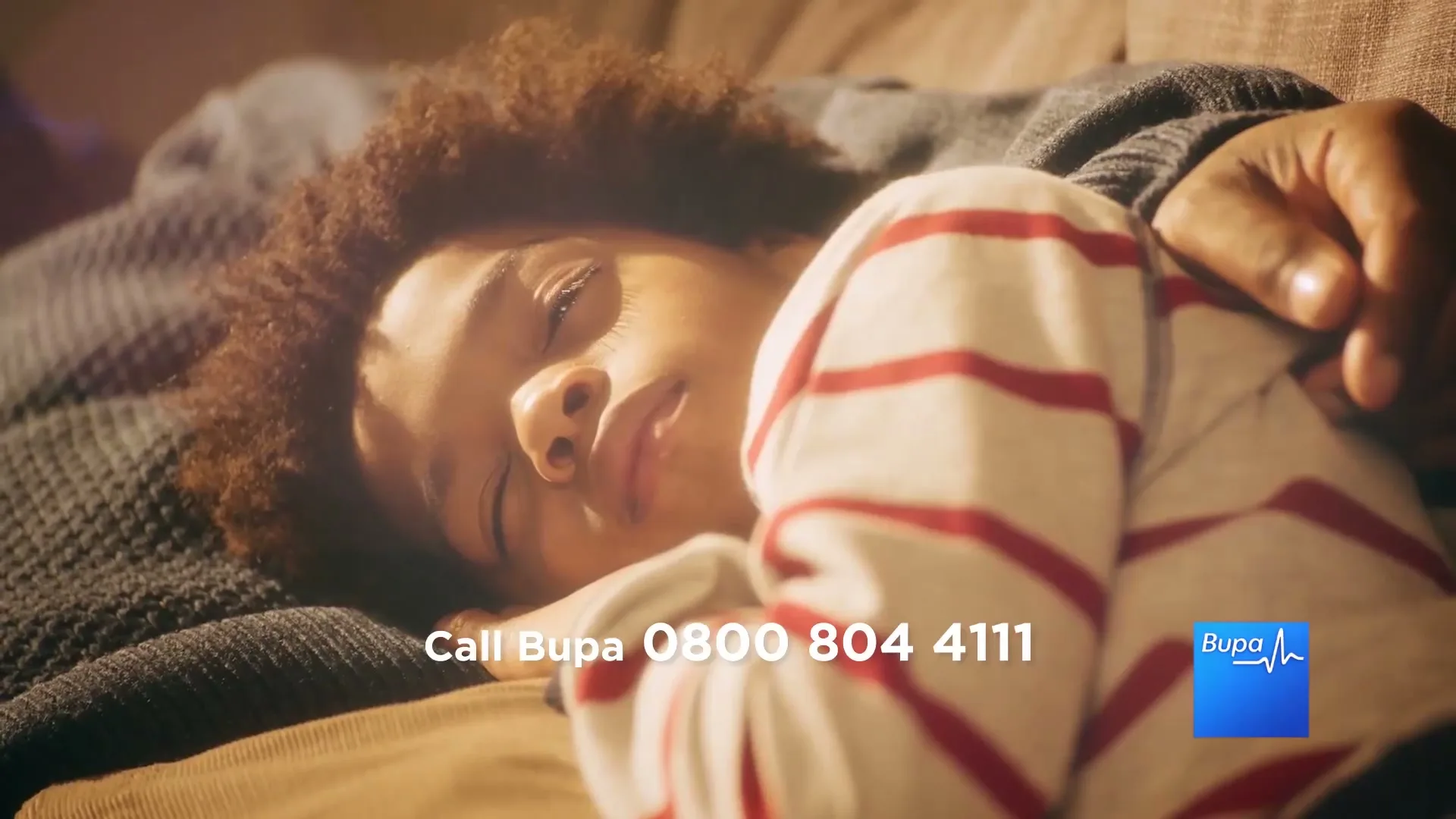 A young child with curly hair sleeps peacefully on a couch, with an adult's arm gently resting on them, in a warm, sunlit living room.