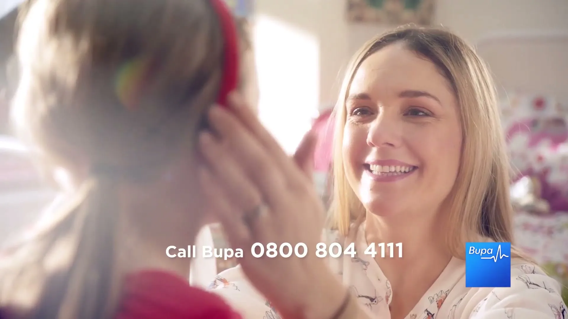 A smiling woman with blonde hair gently touches a young girl's ear, who is wearing red headphones, in a brightly lit bedroom.