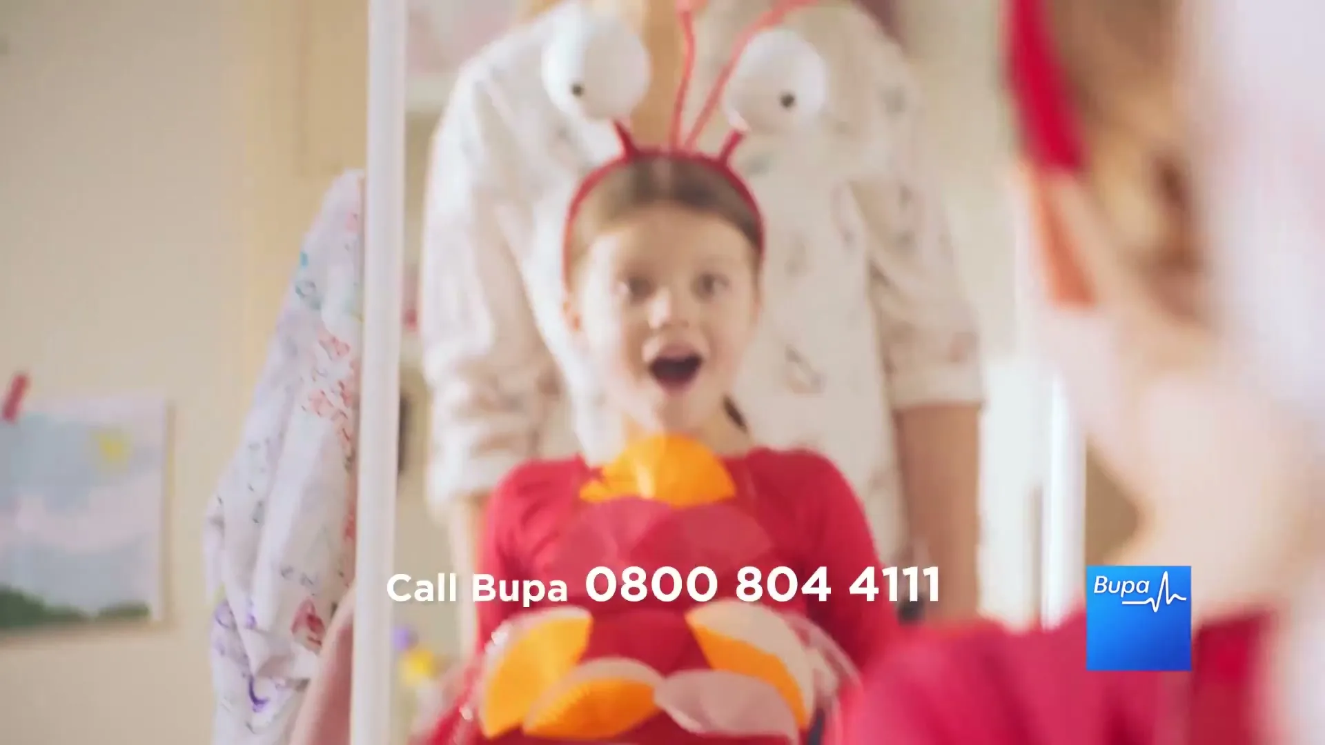 A young girl in a red crab costume with googly eyes looks excitedly at her reflection in a mirror, with an adult out of focus behind her.