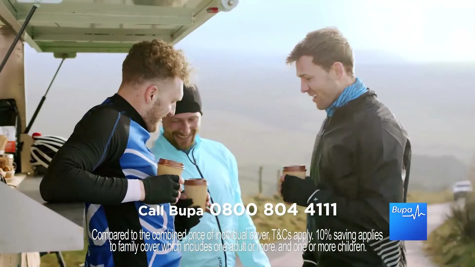 Three men in athletic wear, two cyclists and one runner, laugh and drink coffee from a food truck on a sunny day in a rural landscape.