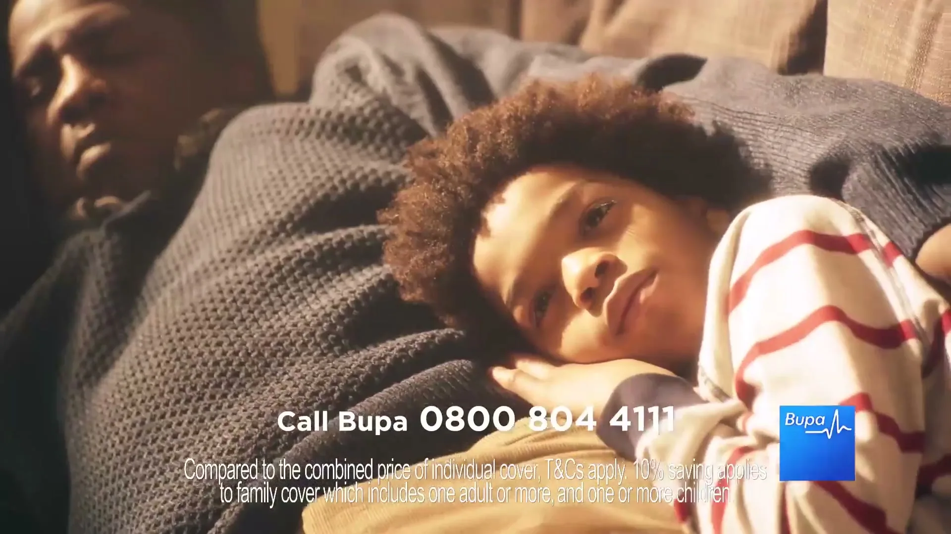 A young boy with curly hair lies on a man's chest, both resting on a sofa in a cozy interior setting.
