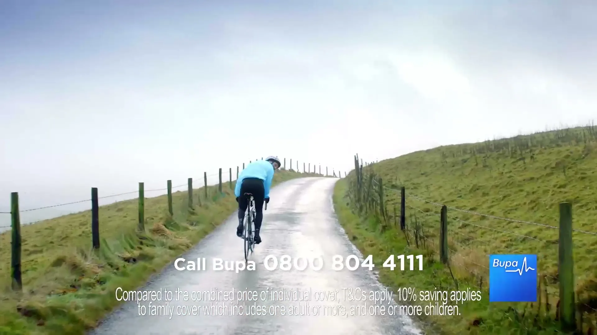 A person in a blue jacket cycles away from the camera up a winding, wet country road lined with fences and green hills under a bright, overcast sky.