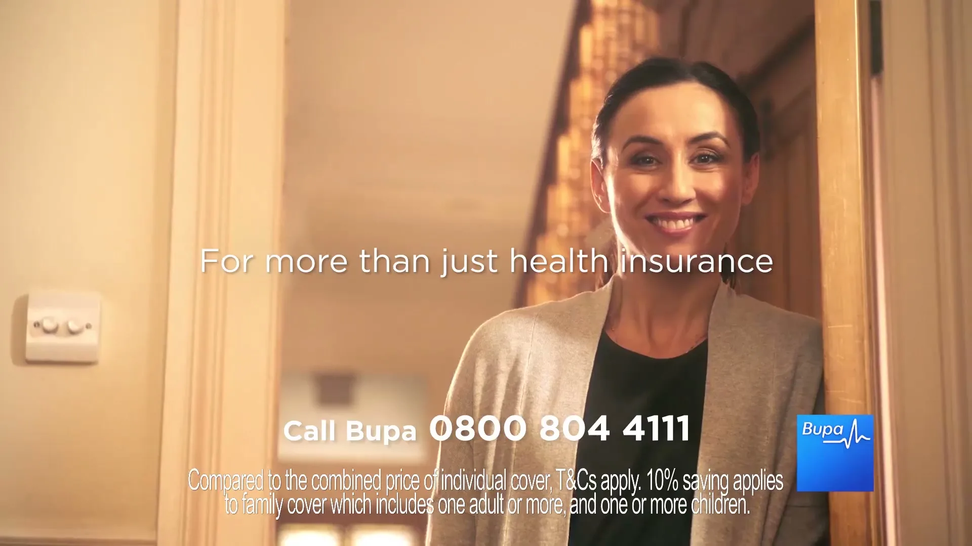 A smiling woman in her 30s stands in a doorway, looking directly at the camera in a warm, inviting interior setting.