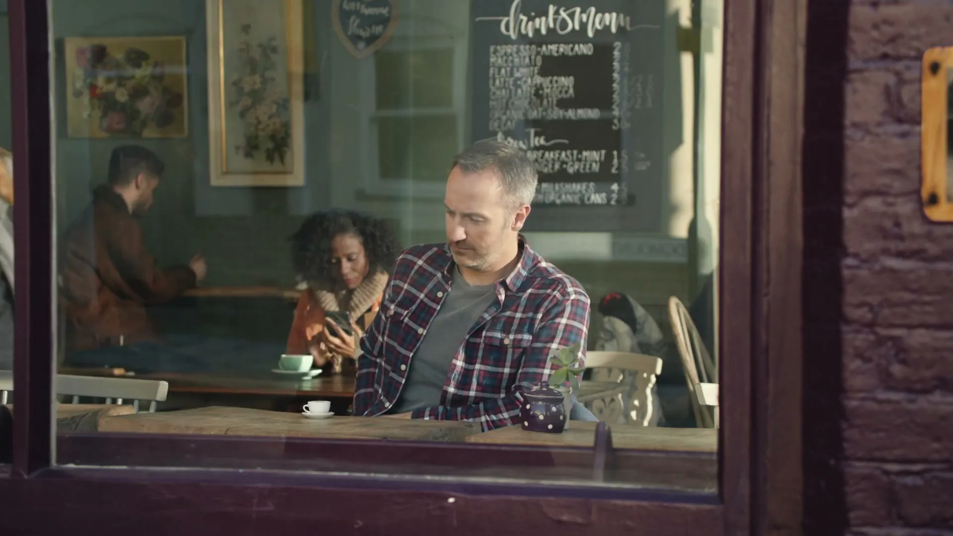 A barista serves a man a small cup of coffee at a wooden table in a cafe, seen through a window.