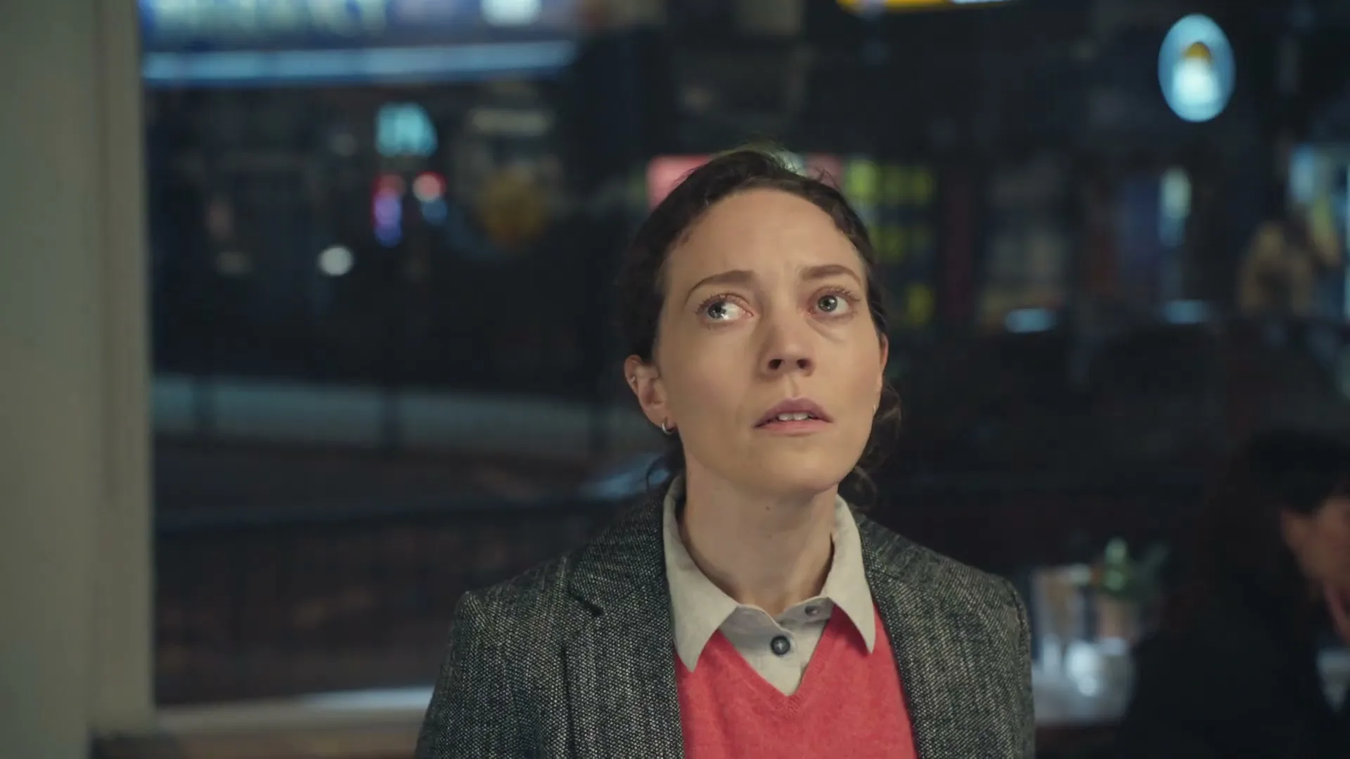 A woman with dark hair, wearing a grey blazer and coral sweater, looks up with a concerned expression inside a restaurant at night.