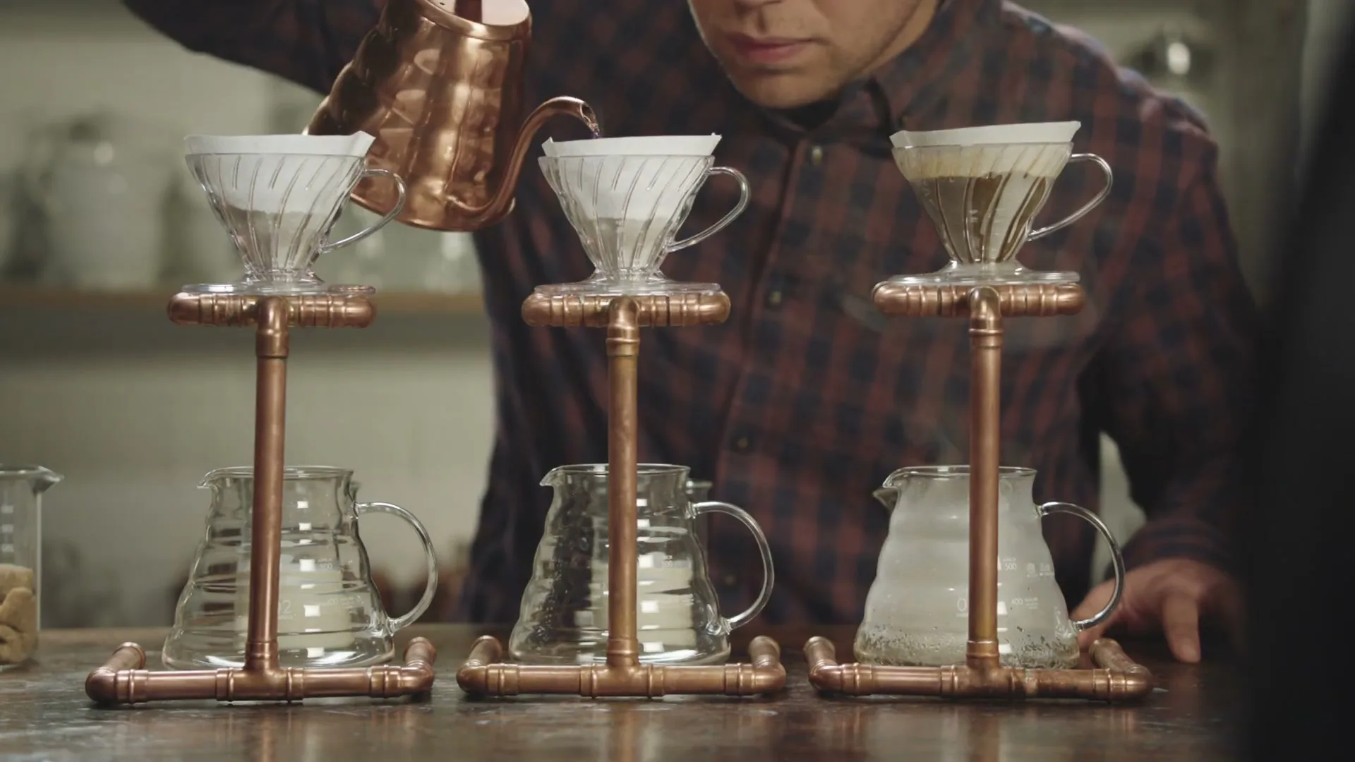 A man in a plaid shirt carefully pours hot water from a copper kettle into a coffee filter, part of a three-station pour-over setup on a wooden counter.