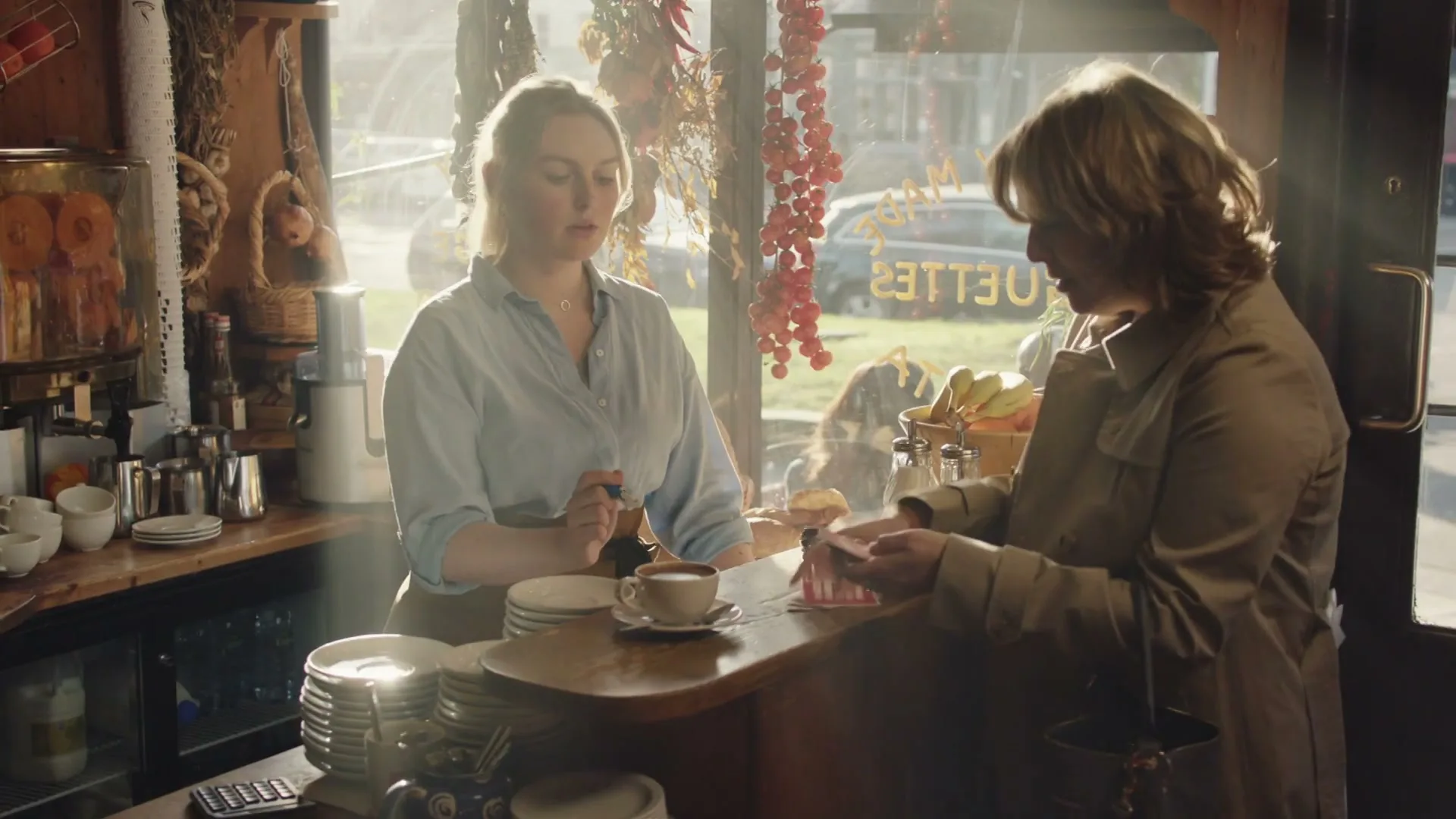 A female barista in a light blue shirt serves a female customer in a trench coat at a sunlit cafe counter.