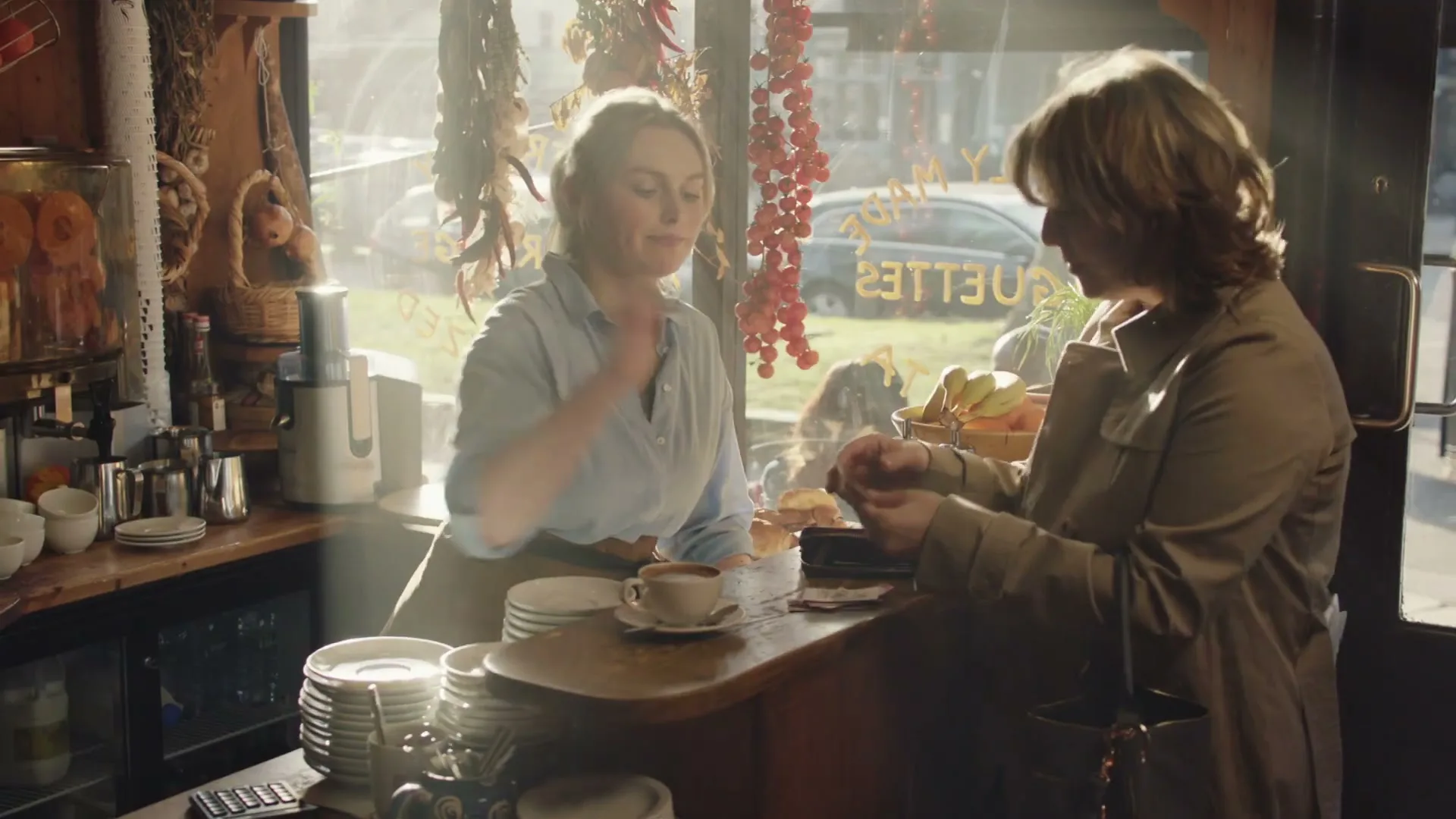 A female barista in a light blue shirt and apron serves a female customer in a trench coat a coffee at a rustic cafe counter during the day.