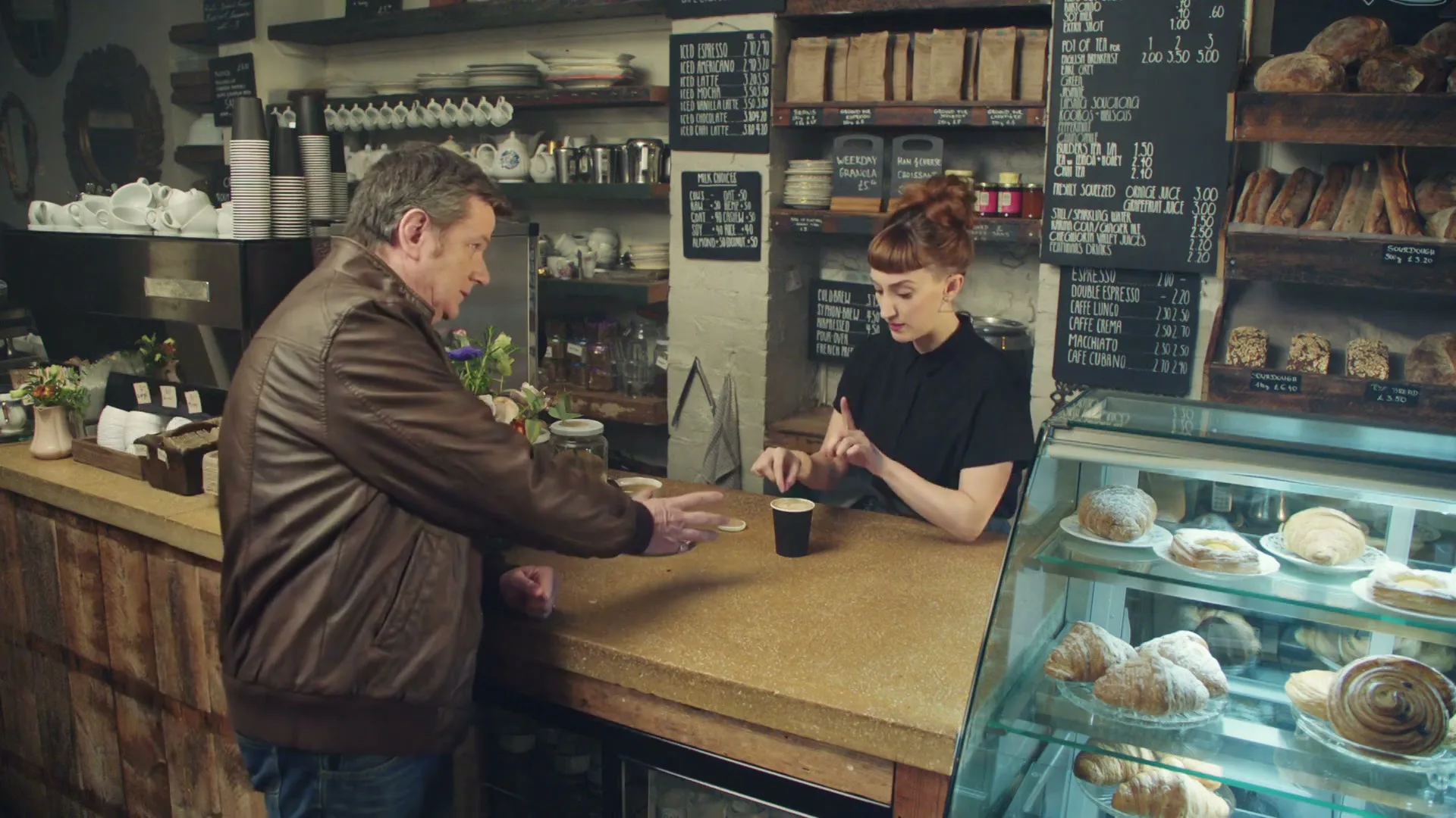 A male customer in a brown leather jacket stands at a coffee shop counter, looking down at a small cup while a female barista prepares a drink.