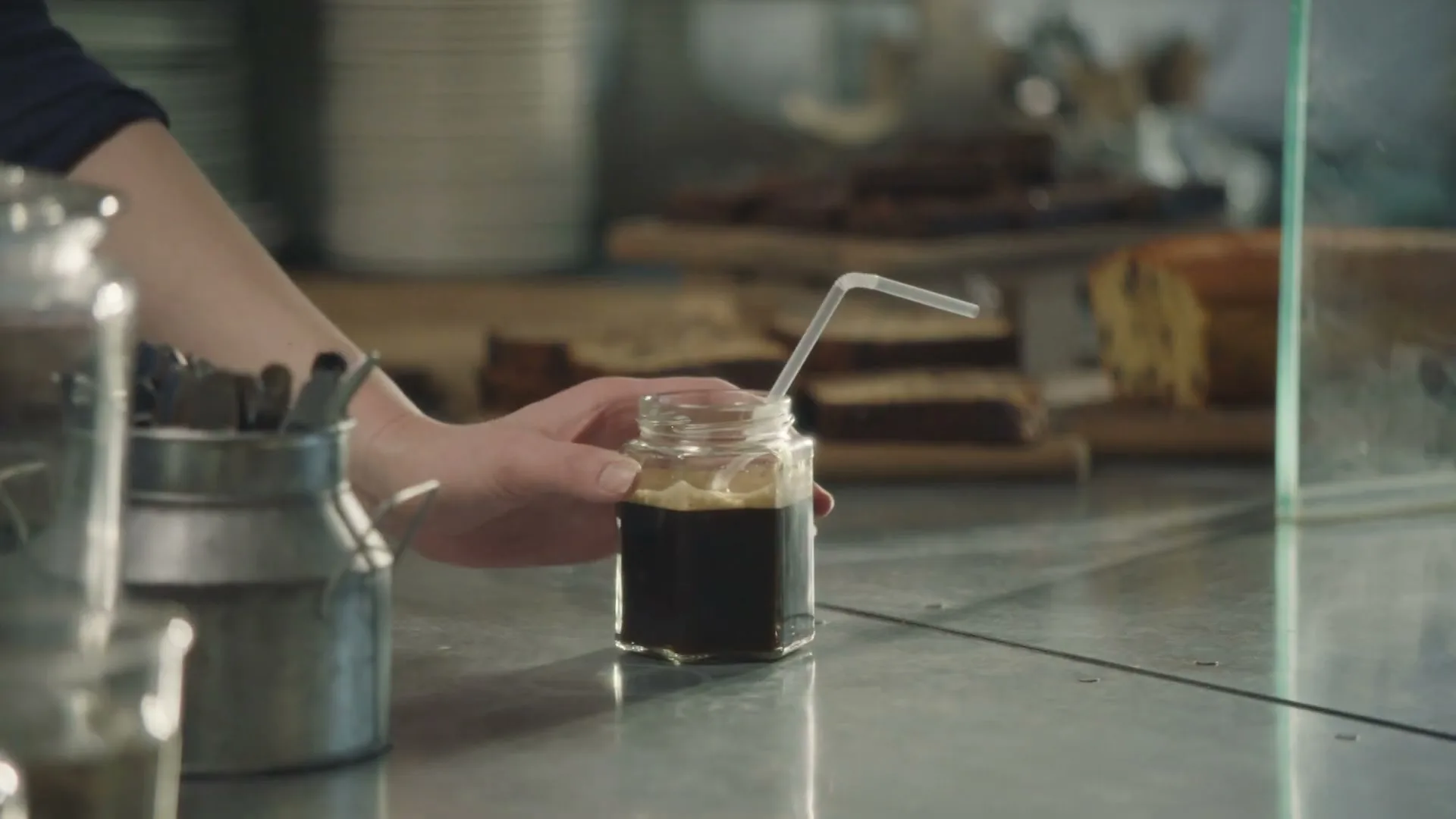 A barista's hand places a small glass jar of iced coffee with a straw on a metal counter in a cafe.