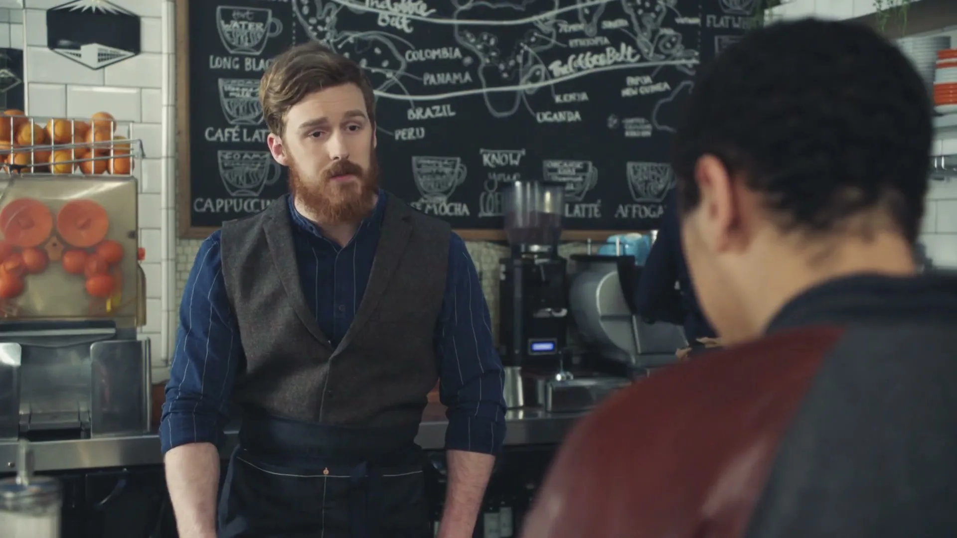 A male barista with a beard looks up and speaks to a customer, who is seen from behind, in a coffee shop.