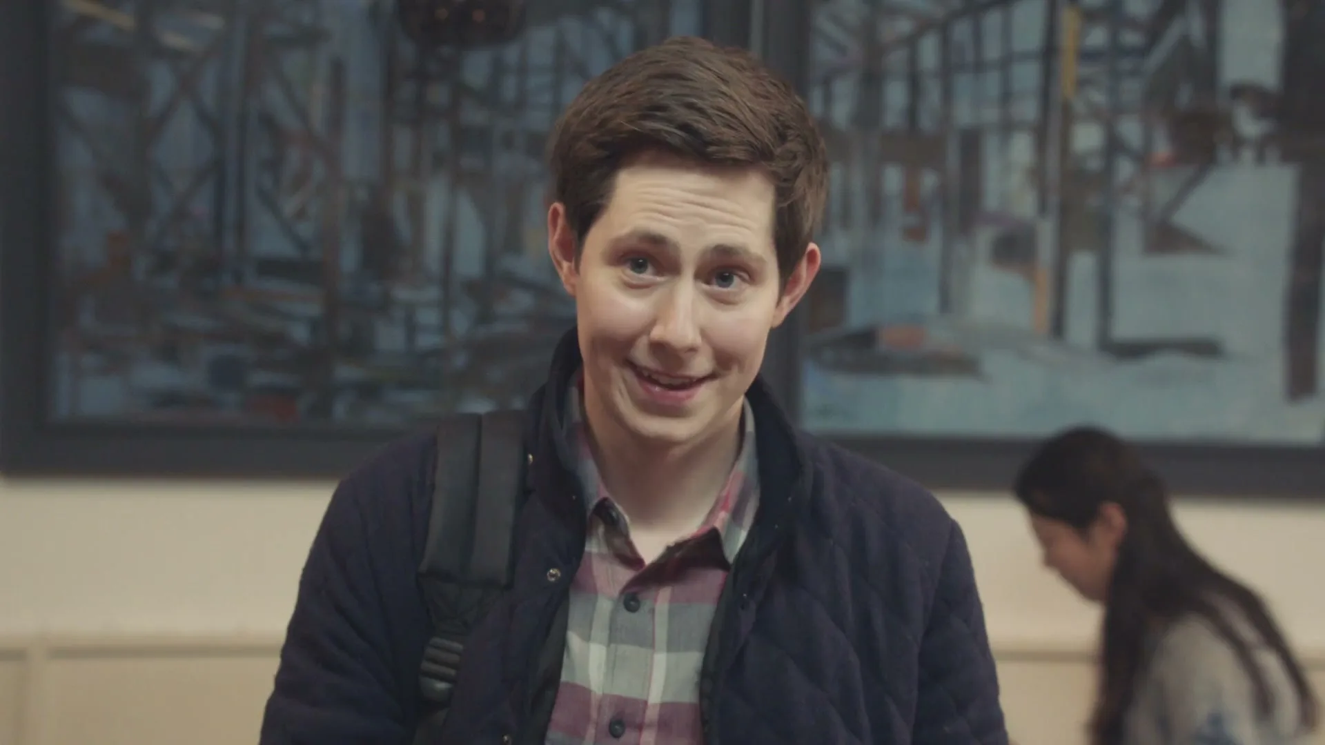 A young man with a backpack smiles awkwardly at the camera in an interior setting, with a blurred painting and another person in the background.