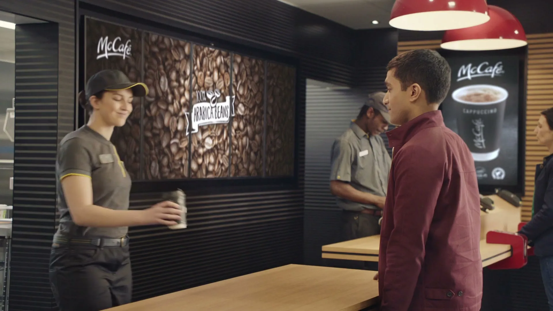 A female McDonald's employee in uniform hands a coffee to a male customer in a red jacket at a McCafe counter.
