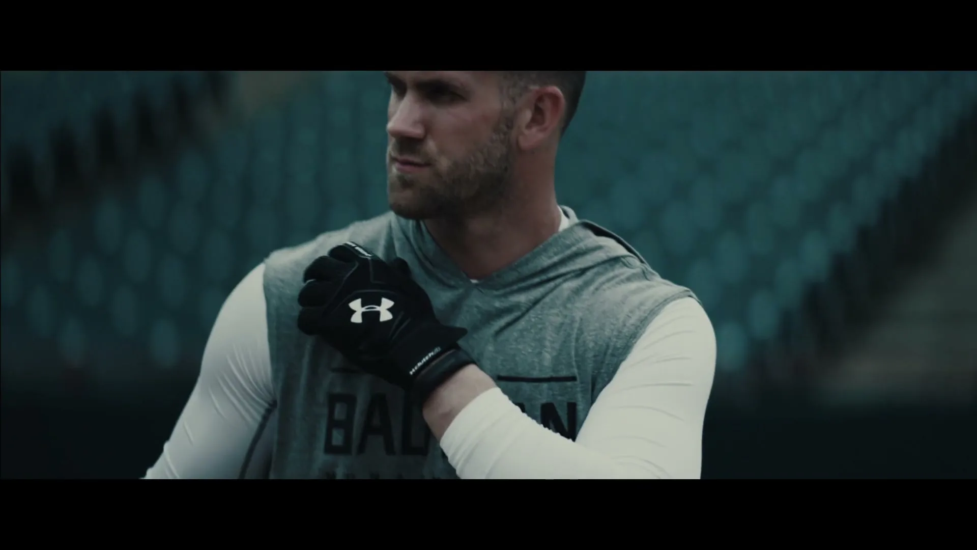 A male athlete in a grey hoodie and white long-sleeve shirt adjusts his black batting glove, looking determined on a baseball field.