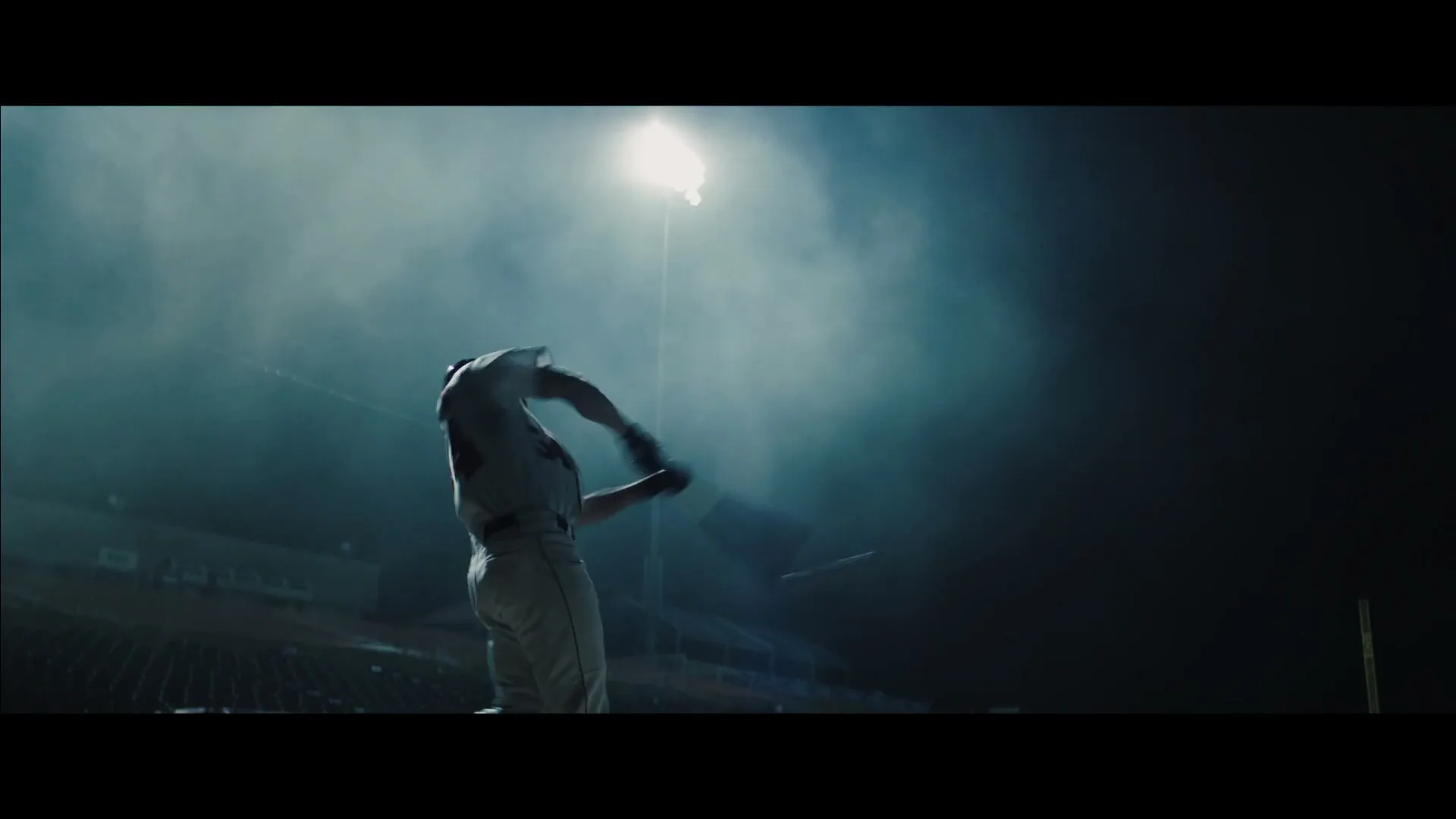 A baseball player in uniform swings a bat at night in a stadium, silhouetted against a bright stadium light.