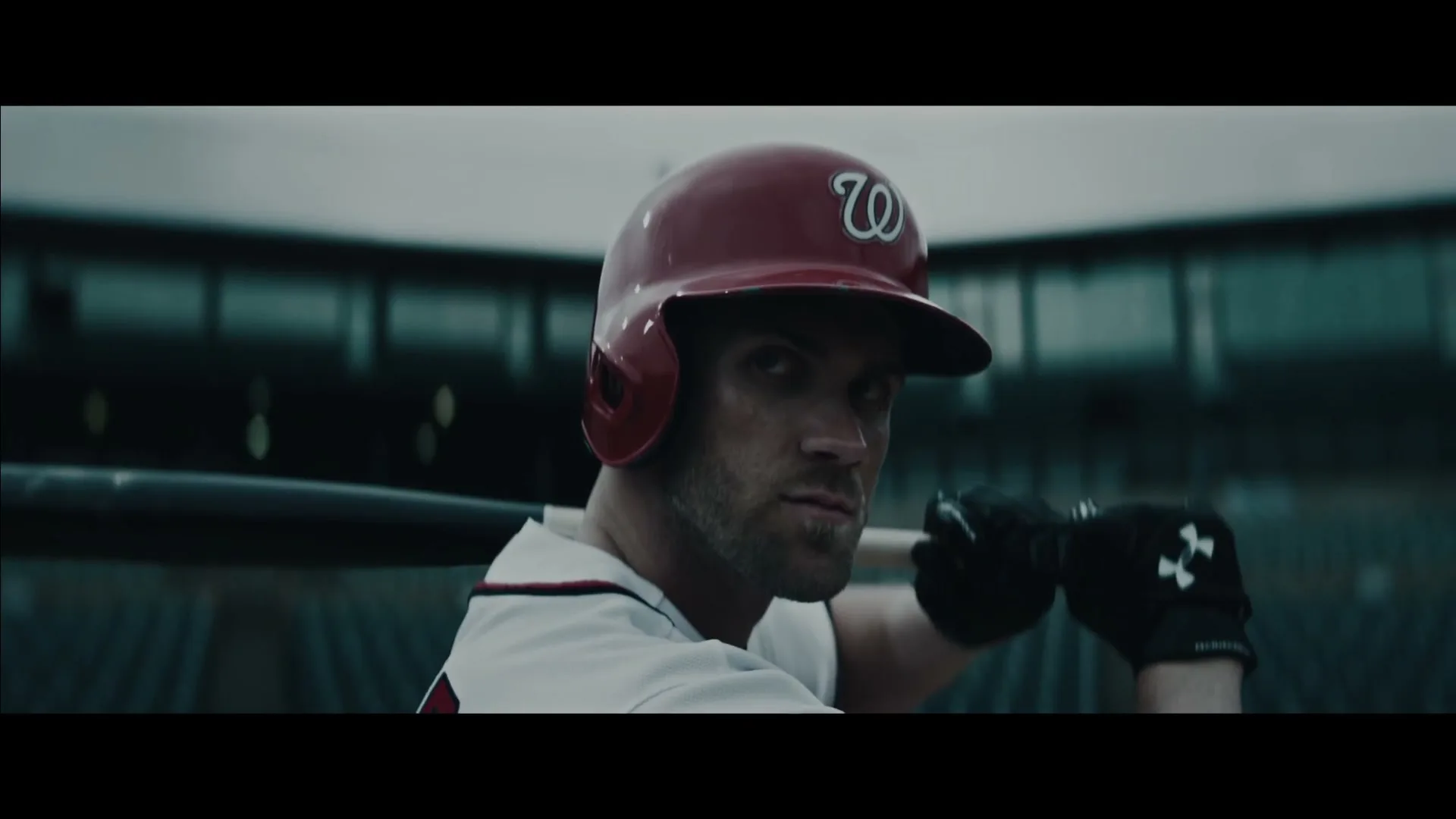 A male baseball player in a red helmet and white jersey holds a bat, looking intensely over his shoulder in a stadium.