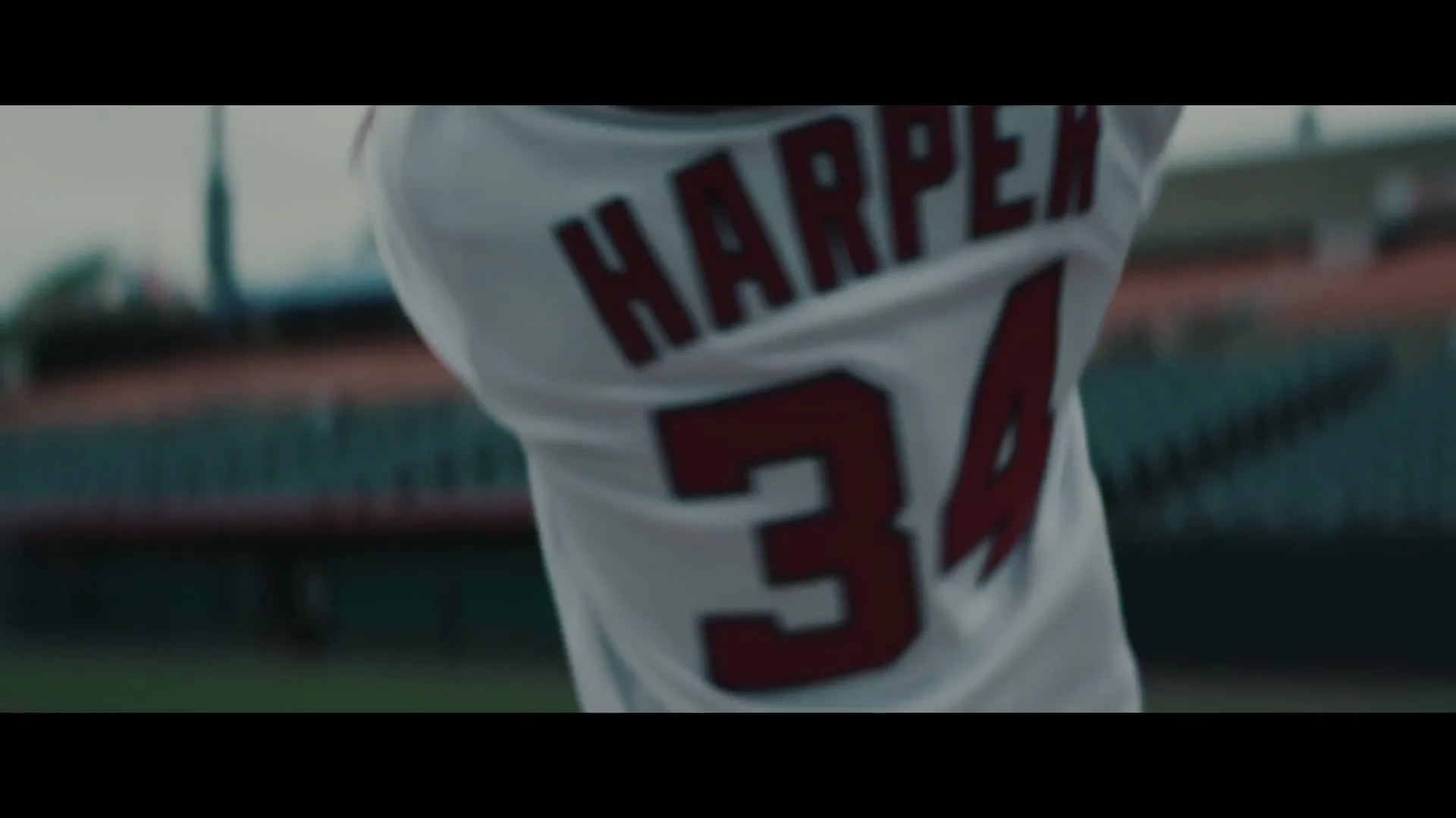 A male baseball player in a white jersey with "HARPER 34" on the back swings a bat in a stadium.