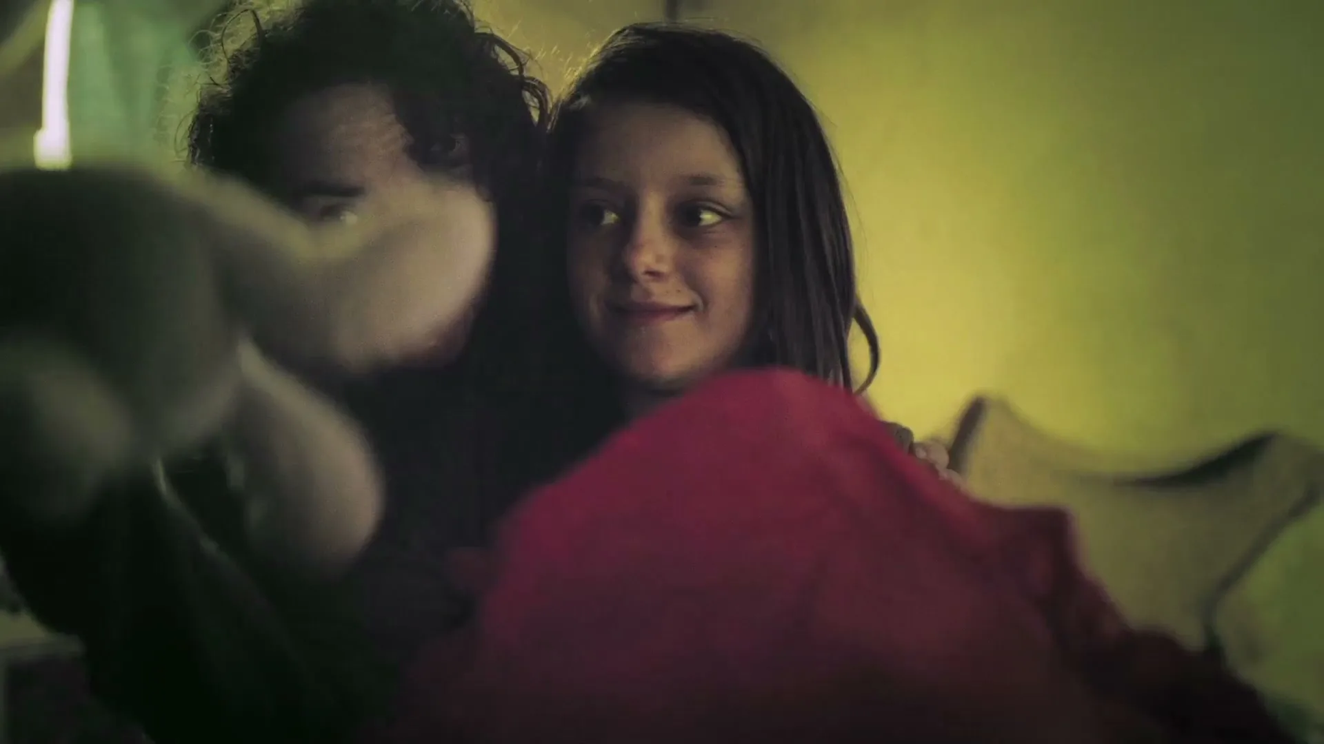 A mother and daughter embrace, smiling as the daughter reveals a white stuffed bunny from a red blanket in a dimly lit room.
