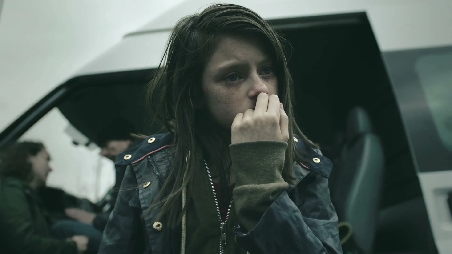 A young girl with dark hair and a worried expression stands in front of an open van door, looking distressed, with other people in the background.