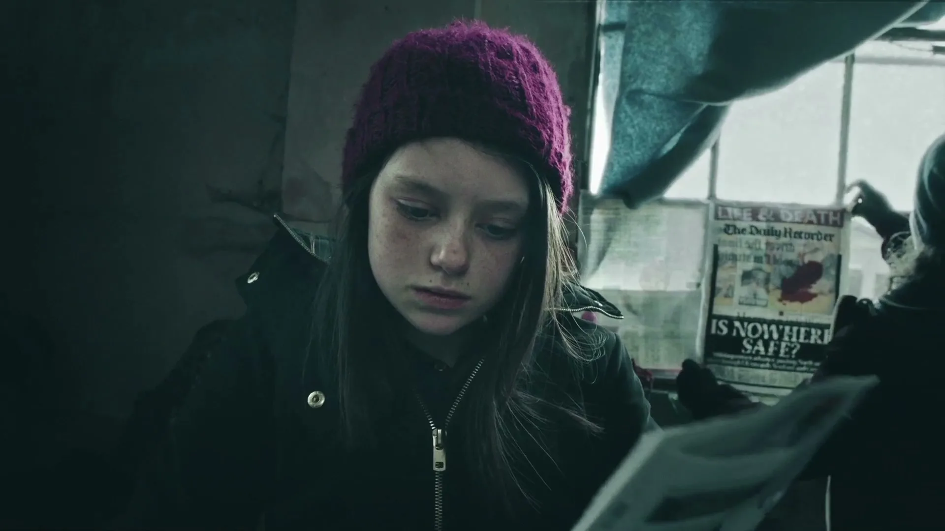 A young girl with a purple beanie looks down sadly while another person holds up a newspaper with a headline about danger in a dimly lit room.