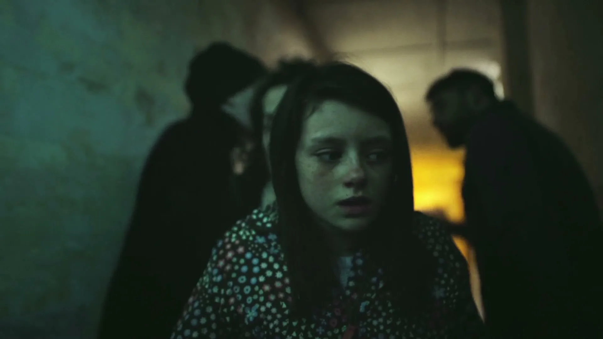 A young girl with dark hair and a floral jacket looks scared, surrounded by adults in a dark, narrow hallway.