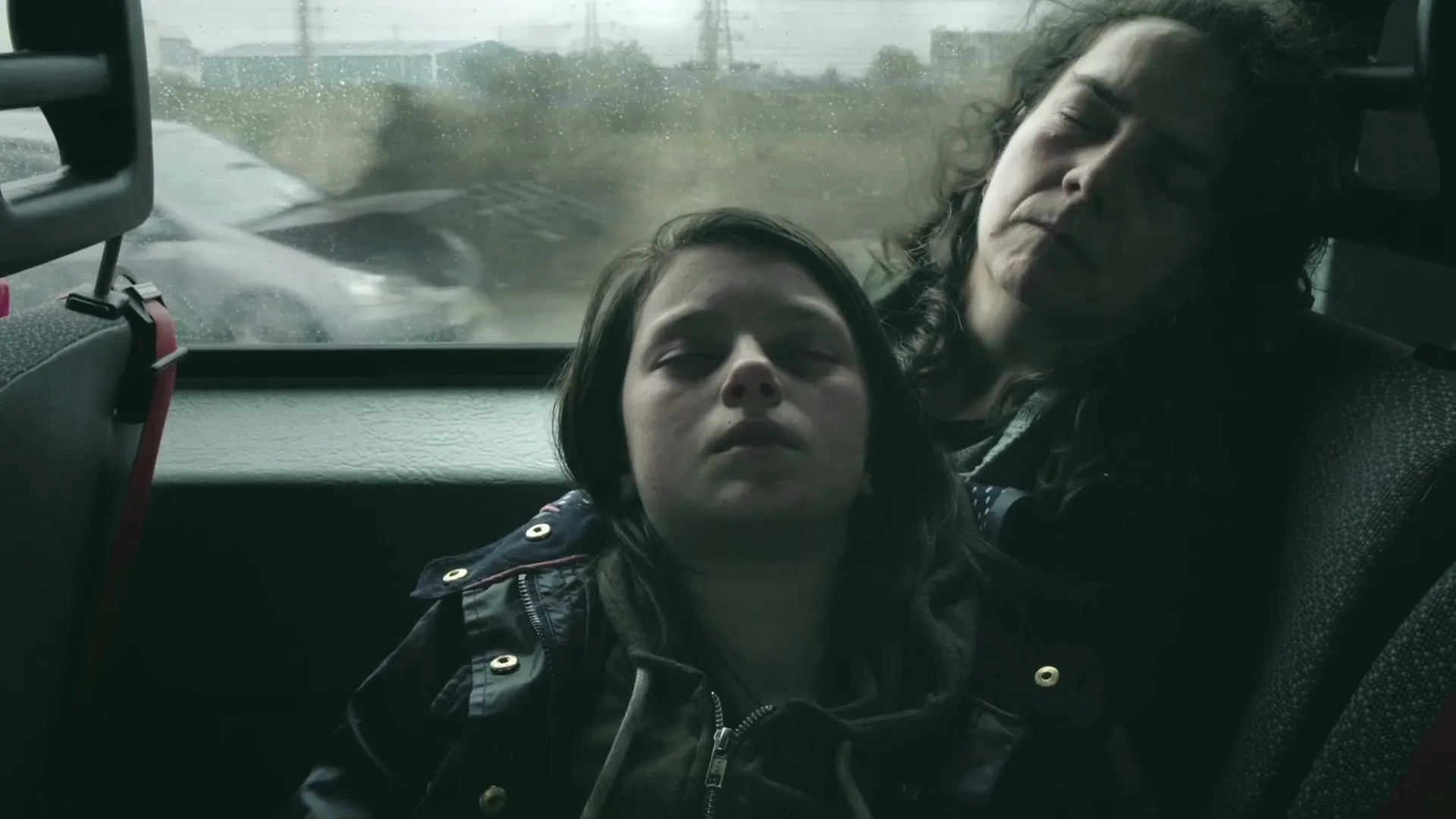 A young girl and an older woman sleep slumped against each other in the back of a moving vehicle, with a blurry, rainy exterior visible through the window.
