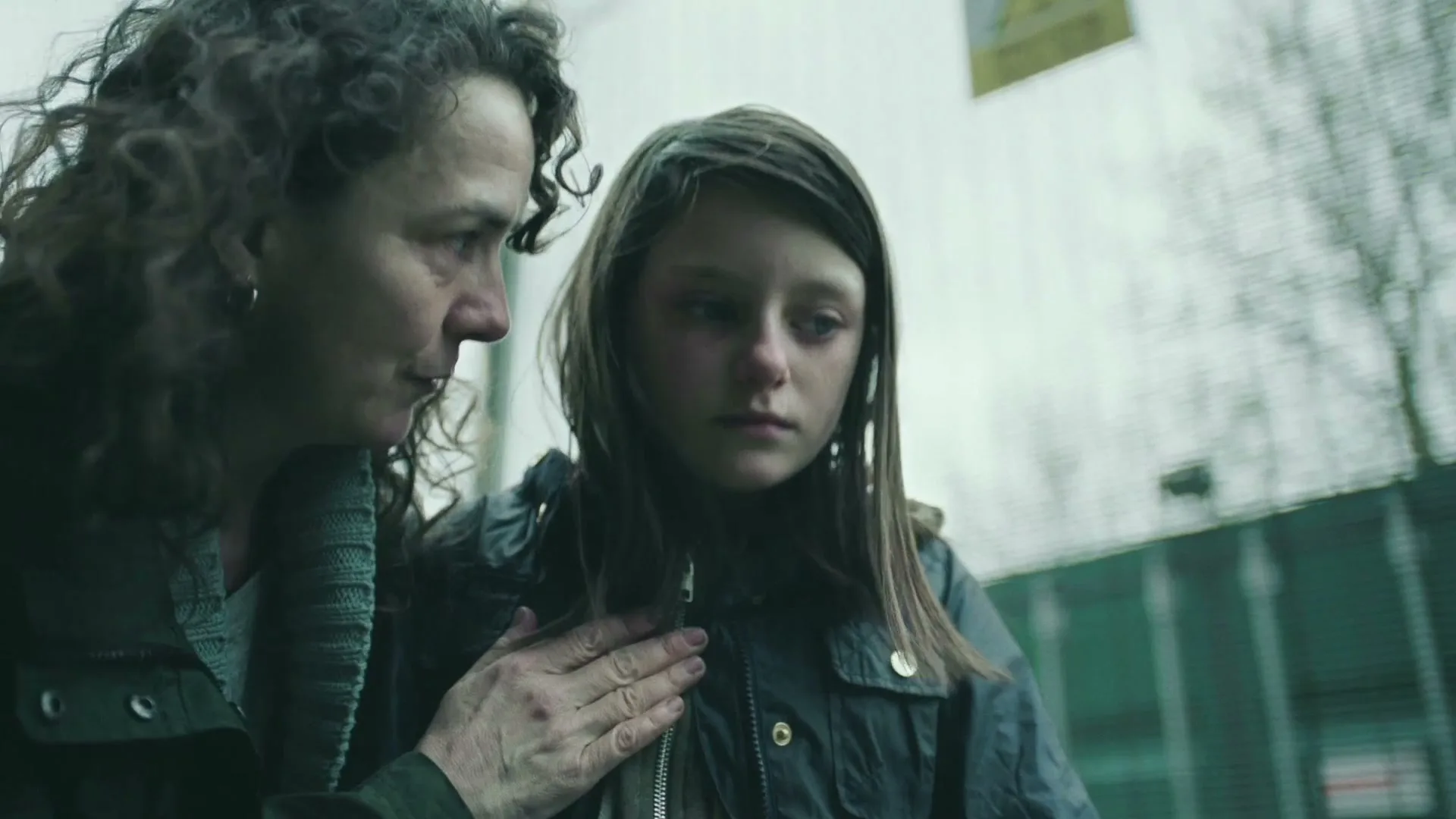 A woman with curly hair comforts a young girl with a bruised face, both looking distressed, outdoors near a fence.