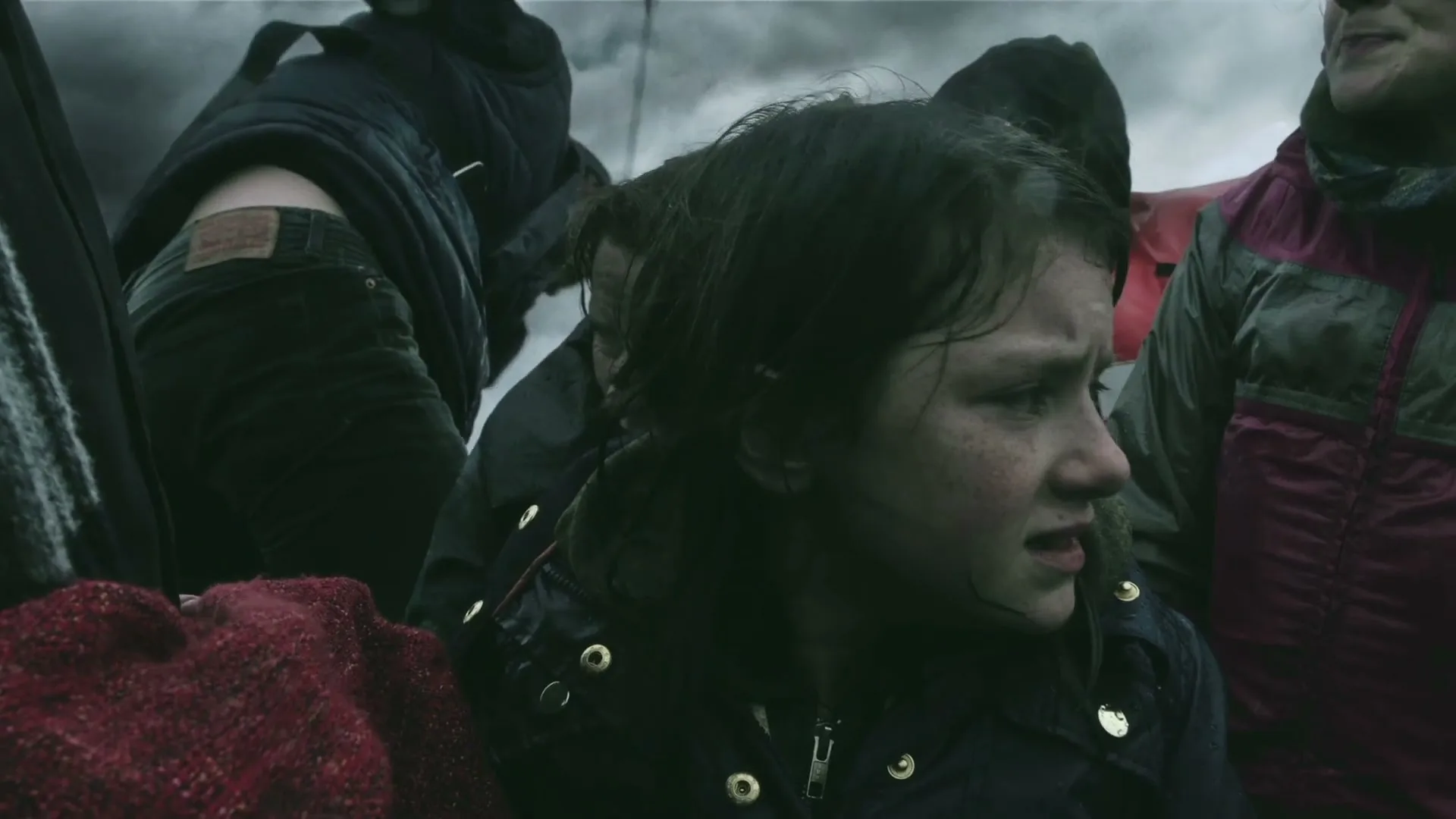 A young girl with a dirty face and wet hair looks distressed while surrounded by other people in a crowded, chaotic setting.