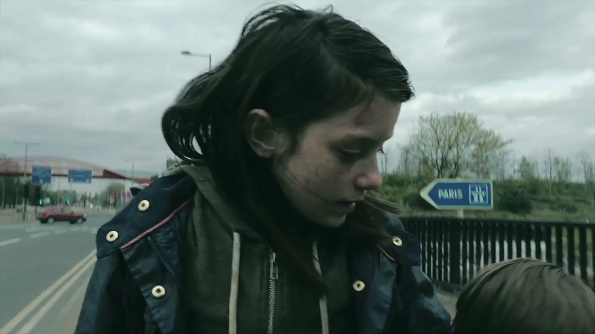A young girl with a dirty face and dark hair looks down, wearing a blue jacket and grey hoodie, on a bridge with a "Paris" sign in the background.