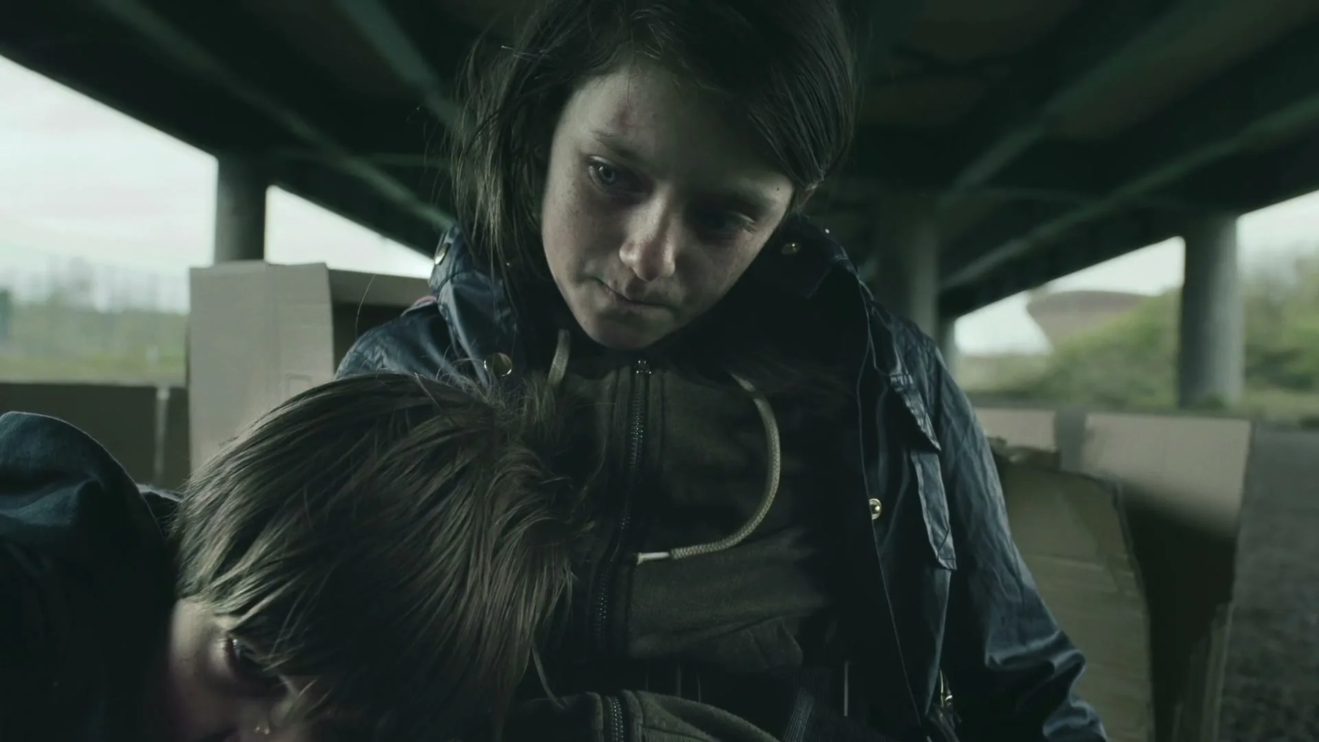 A young girl with a bruised face holds another child, looking down with a somber expression under a bridge.
