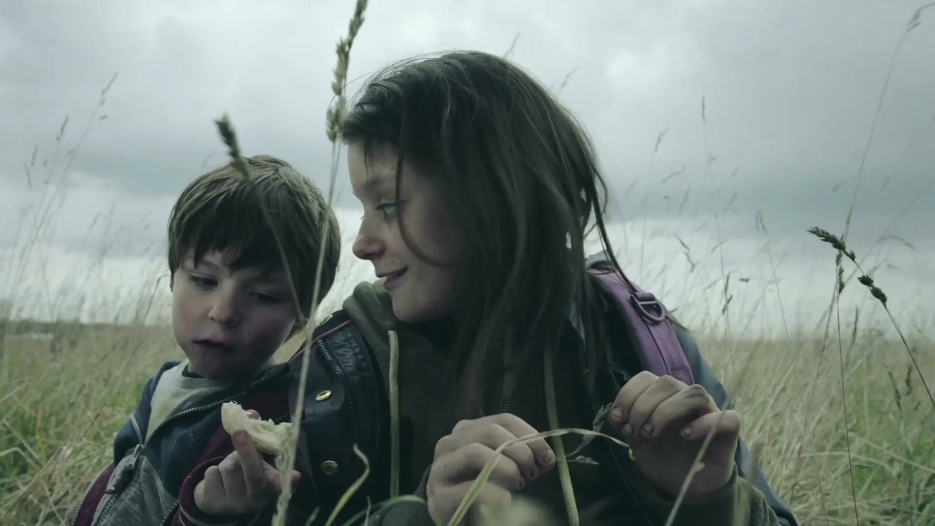 A young girl and a boy sit in a field of tall grass, the girl looking at the boy who holds a piece of bread, under a cloudy sky.