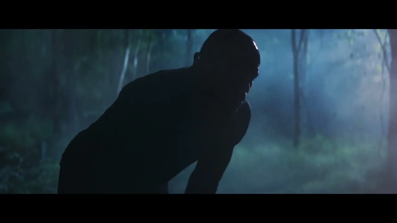 A male athlete, silhouetted against a misty, dark forest, slowly rises and looks up, appearing determined.