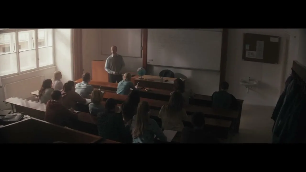 A male teacher stands at the front of a classroom, addressing a group of students seated at wooden desks, with sunlight streaming through a large window.