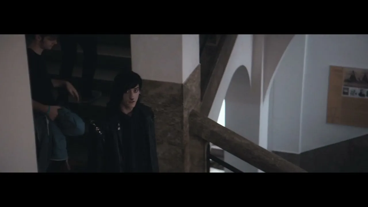 A young man with dark hair and a black jacket stands on a staircase, looking around with a neutral expression in a dimly lit building.