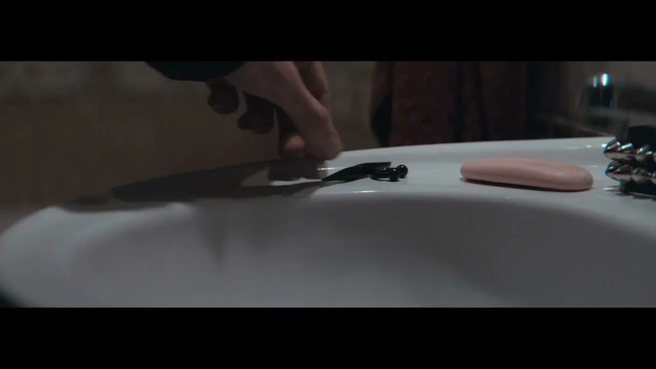 A person's hand places a small, dark object onto the edge of a white bathroom sink, next to a pink bar of soap.